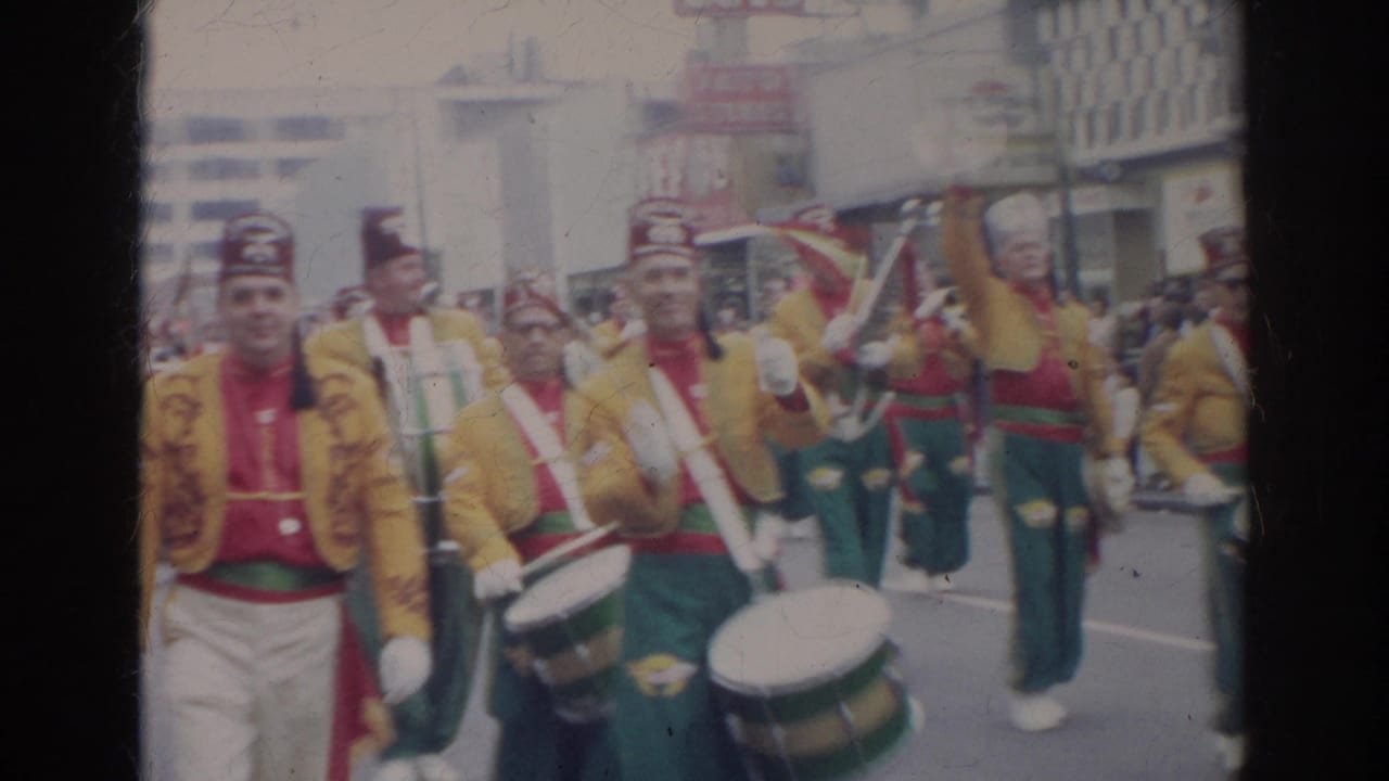 Uniformed parade musicians approach the camera with drums and brass visible in the street.