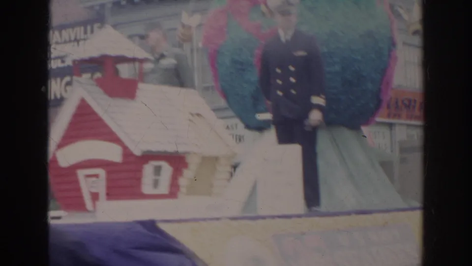 A decorated parade float in Norfolk includes a small red-and-white house prop, a uniformed man, and large colorful float shapes.