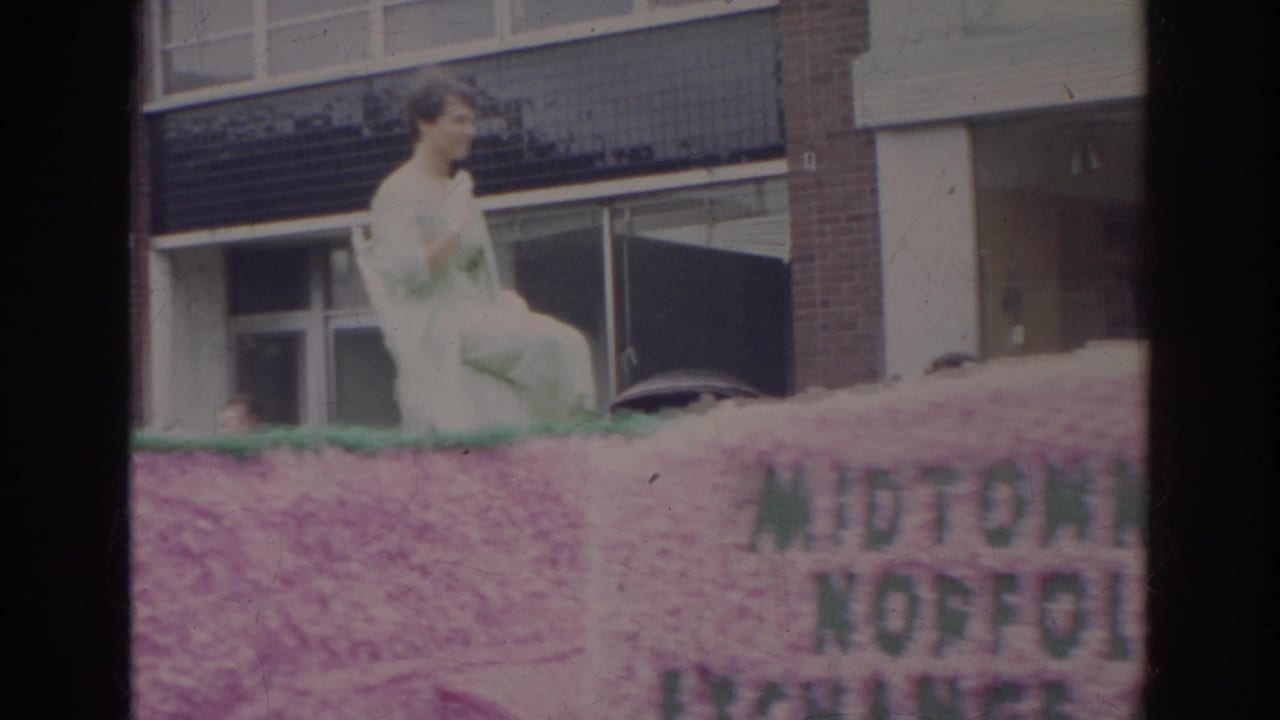 Woman seated on a pink floral parade float passing brick buildings.