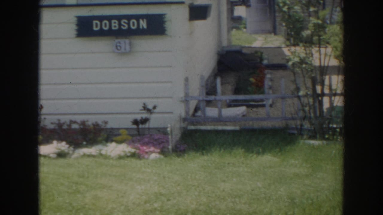 A closer 1966 home-movie view shows the DOBSON sign over number 61, with white siding, lawn, flower beds, and a small side entry area.