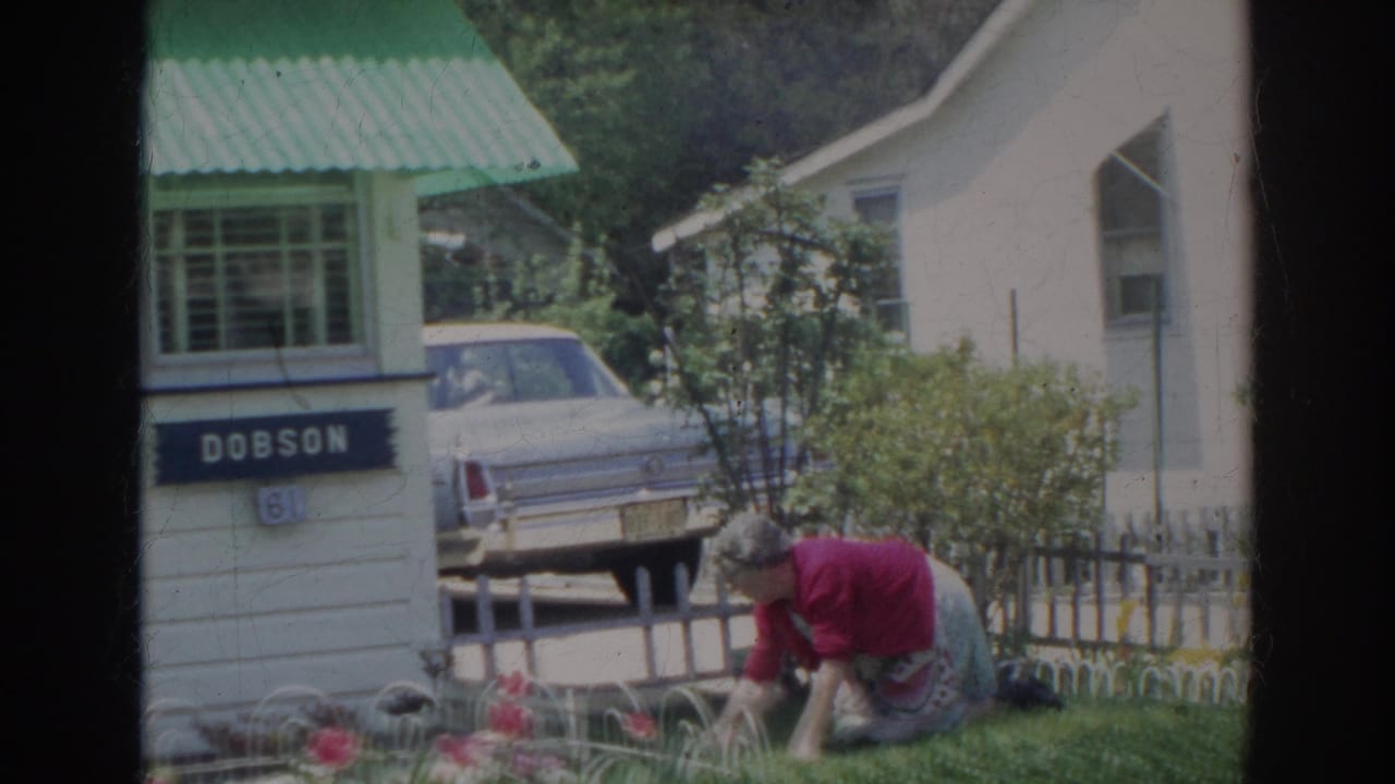 Woman in a red sweater bends over a front garden beside a white house and parked car.