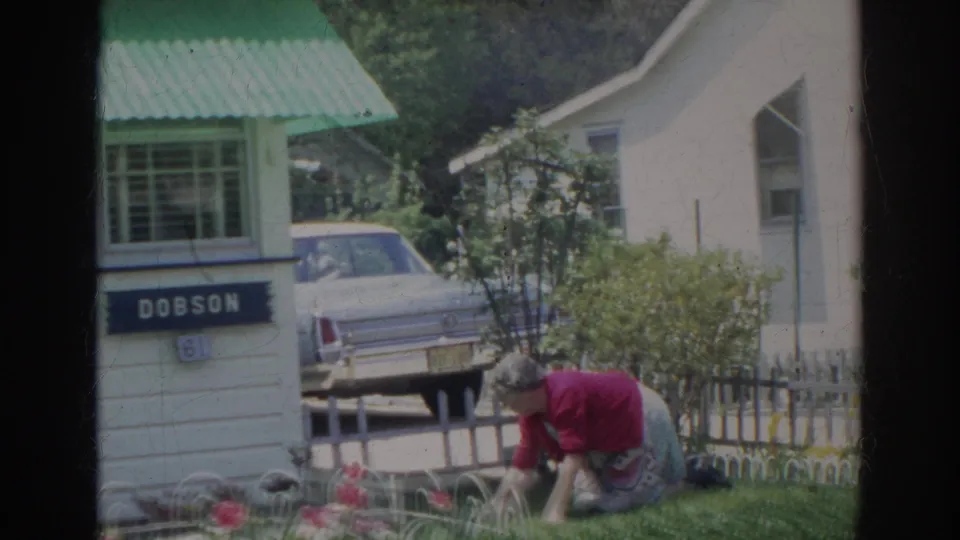 In 1966 Norfolk home-movie footage, a woman in a red sweater works in a flower garden outside a white house with a green awning.