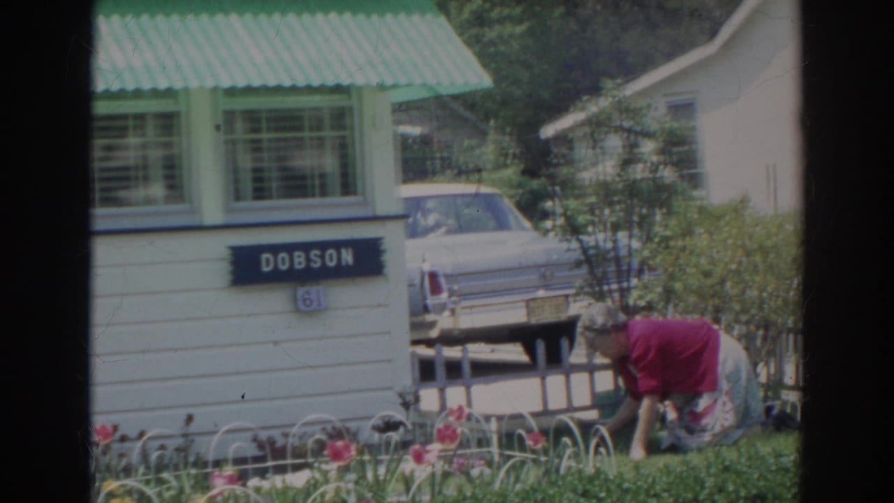 Woman bends near flowers in front of a house marked Dobson, with a parked car nearby.