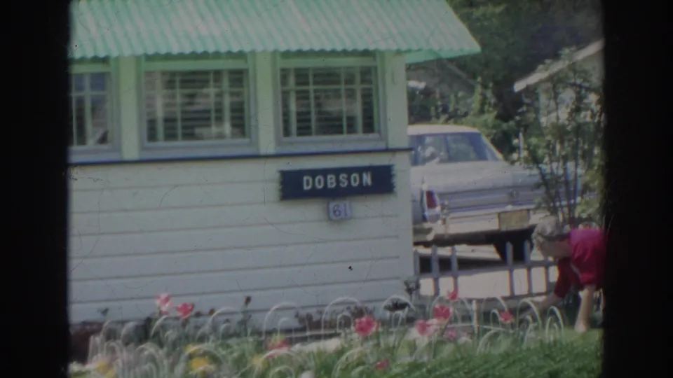 The frame emphasizes the white house, green awning, Dobson sign, flowers, and parked car while the gardener is partly cropped at lower right.