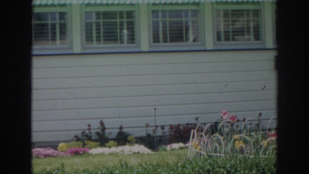 1966 Norfolk home-movie frame of a white house side with green window trim, lawn, flowers, and a low white garden fence.