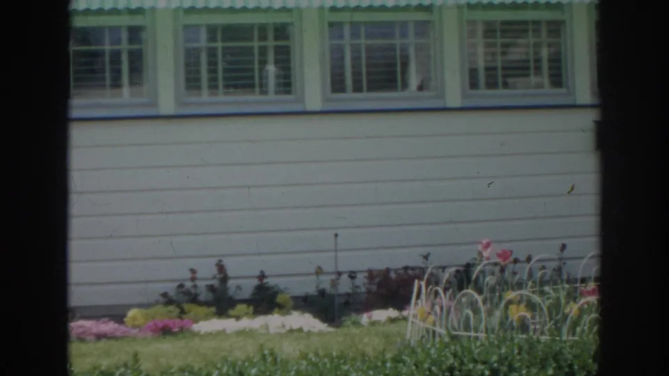 1966 Norfolk home-movie frame of a white house side with green window trim, lawn, flowers, and a low white garden fence.