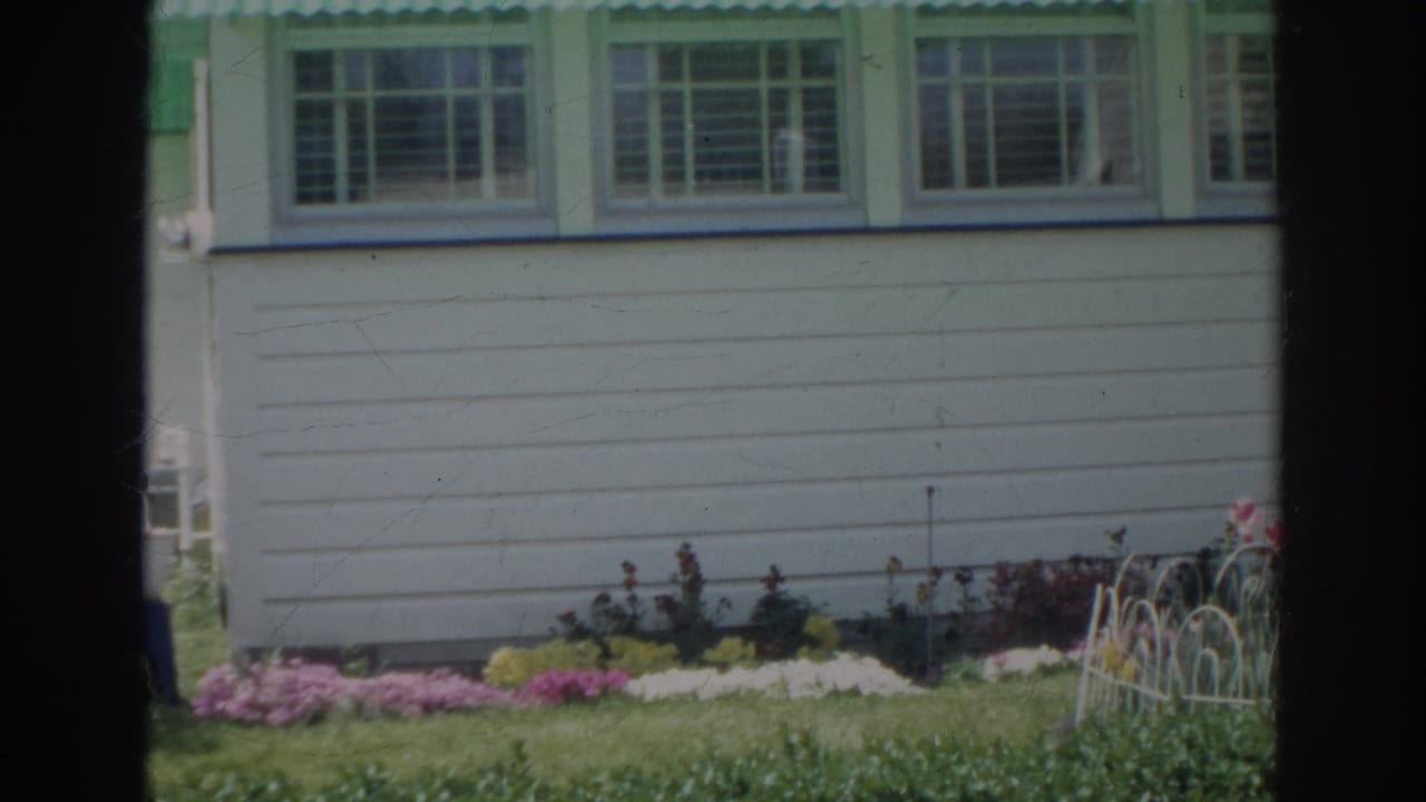 1966 Norfolk home-movie frame centered lower on the clapboard wall, with flower beds, lawn, and white metal garden edging.