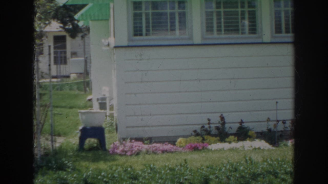 1966 Norfolk home-movie frame showing the side of a white clapboard house, green-trimmed window, lawn, flower bed, and yard fixture.
