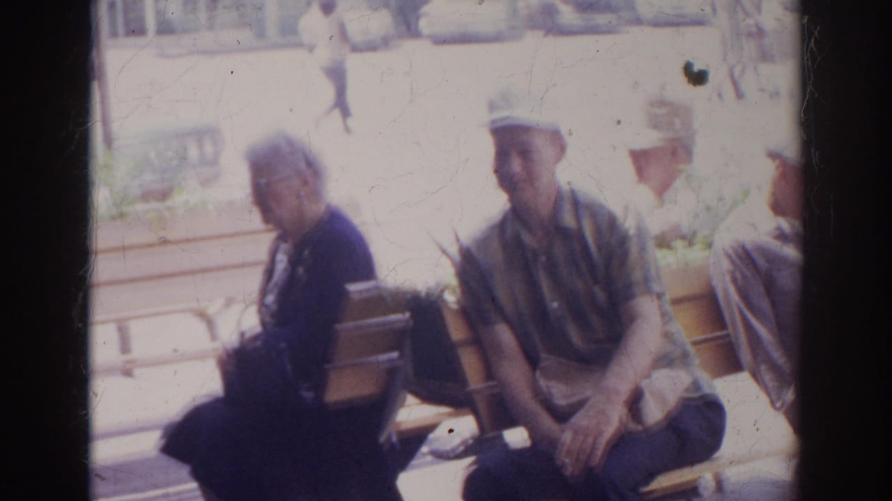 In 1966 Norfolk, an older woman in dark clothing and a man in a white hat sit on a wooden bench in bright sun.