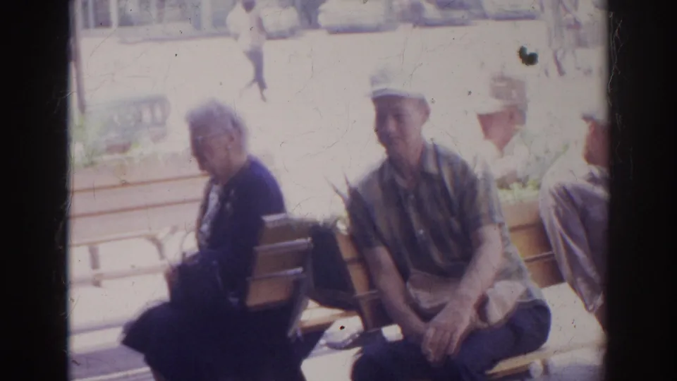In 1966 Norfolk, an older woman in dark clothing and a man in a white hat sit on a wooden bench in bright sun.