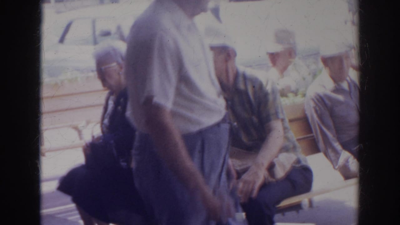 A man passes close to the camera while older adults remain seated on a bench.