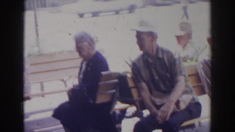 Older adults sit side by side on a wooden bench, with another seated figure partly visible in the bright 1966 Norfolk setting.