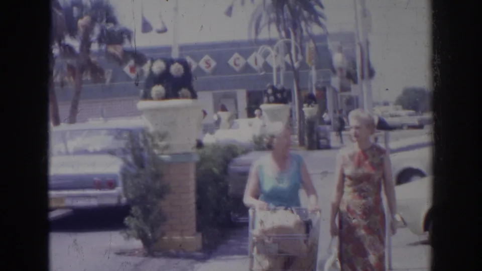 In Norfolk, Virginia, 1966, two elderly women walk toward the camera with groceries near parked cars and supermarket signage.
