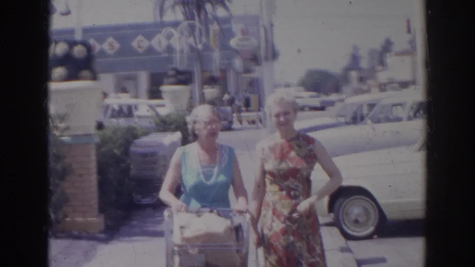 Two elderly women in sleeveless dresses pause or continue forward with groceries in a Norfolk supermarket parking lot in 1966.