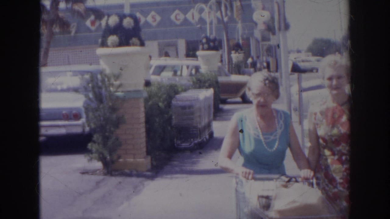 The women pass near the camera with groceries, with mid-century cars and supermarket decoration visible behind them in Norfolk, 1966.