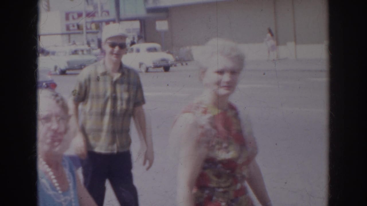 A floral-dressed woman passes close to the lens as a man in sunglasses crosses the Norfolk supermarket parking area in 1966.