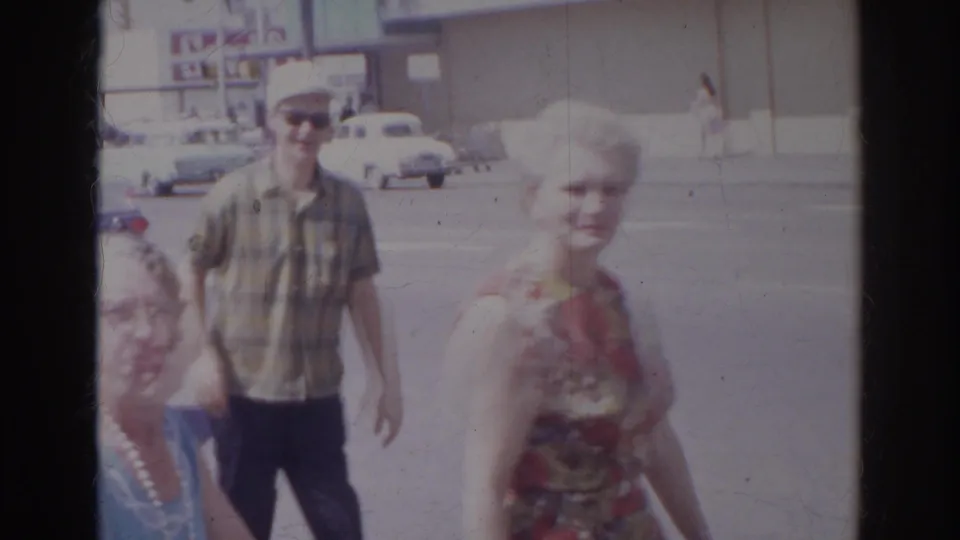 A floral-dressed woman passes close to the lens as a man in sunglasses crosses the Norfolk supermarket parking area in 1966.