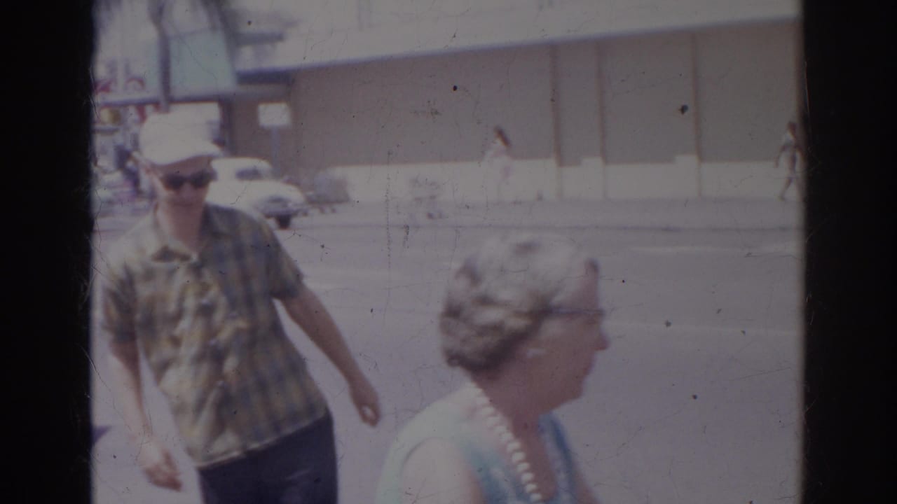 In 1966 Norfolk, Virginia, an older man in a plaid shirt and white cap walks near an older woman beside a street with cars and storefronts.