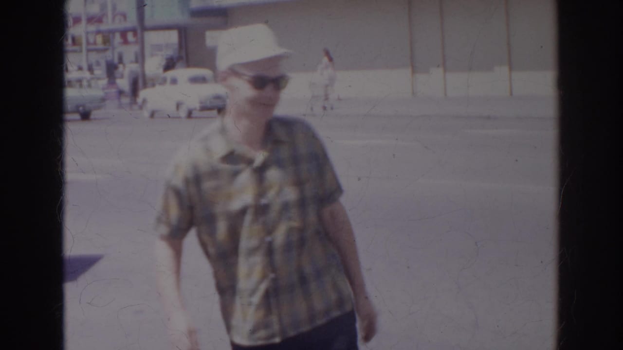 Man in plaid shirt and white cap walks toward the camera on a bright street.