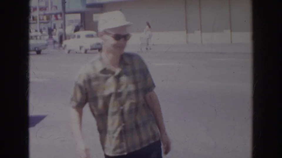 A man in a plaid shirt, white cap, and sunglasses approaches the camera on a bright Norfolk street with vintage cars behind him.