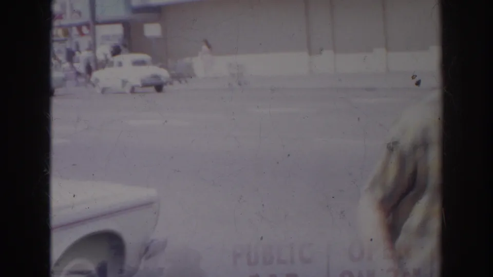 The man passes out of frame in a handheld 8mm view of a Norfolk street, with parked cars and commercial buildings still visible.