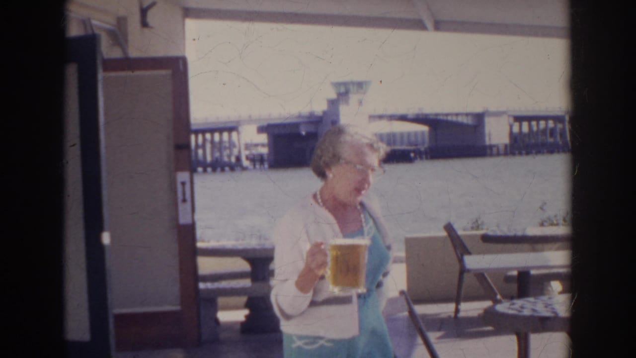 1966 Norfolk home-movie frame of a woman in light clothing holding beer on a waterfront patio with seating and a bridge behind her.