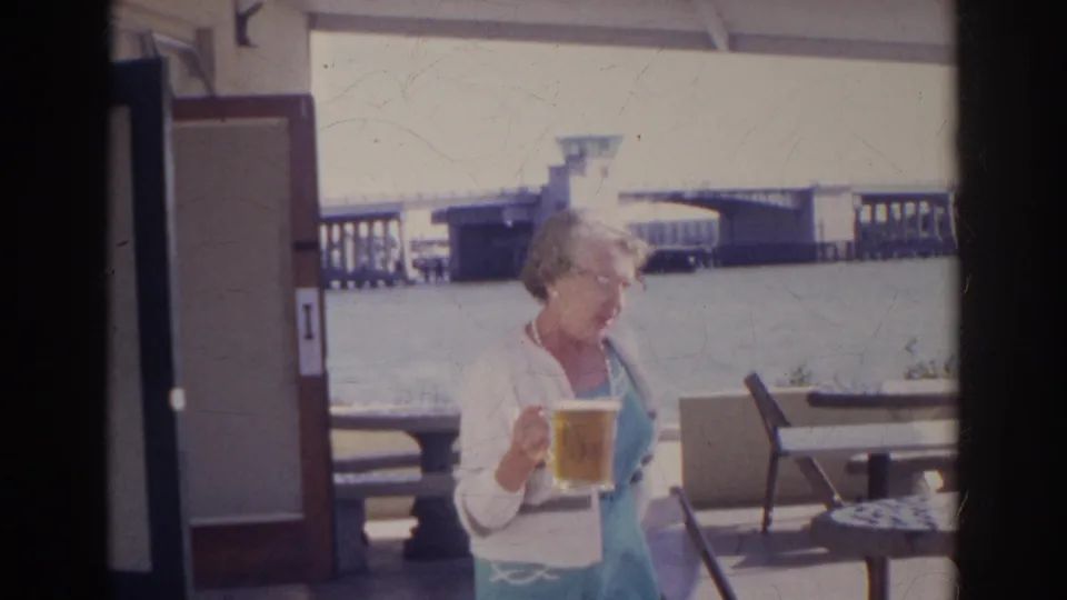 1966 Norfolk home-movie frame of a woman in light clothing holding beer on a waterfront patio with seating and a bridge behind her.