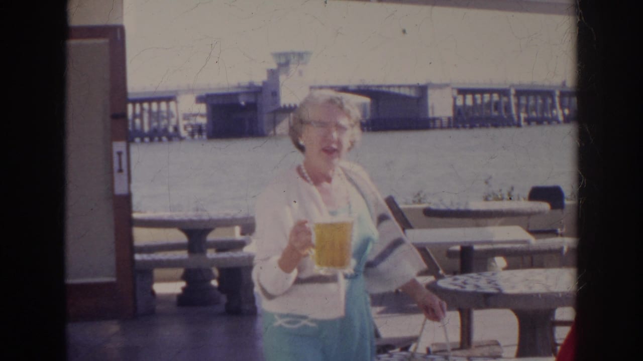 1966 Norfolk frame showing a woman carrying beer and a bag beside patio tables, with waterfront railings and a bridge in the background.