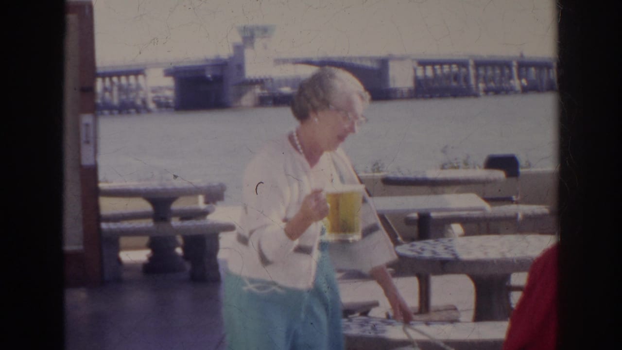 In Norfolk, Virginia, 1966, a woman in a white cardigan and turquoise skirt carries a large beer glass beside stone picnic tables by the water.