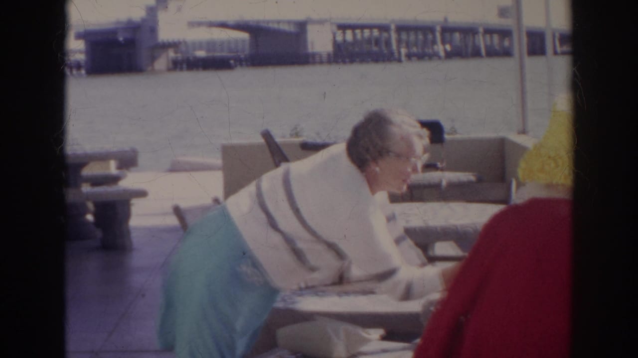 At the waterfront picnic table, the woman bends forward while a seated companion in red and a yellow hat is visible in the foreground.