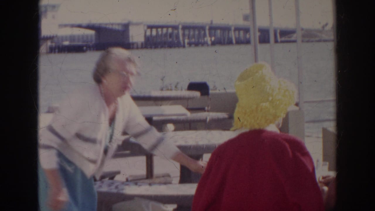 Adults at an outdoor waterfront cafe with stone tables and a pier in the background.