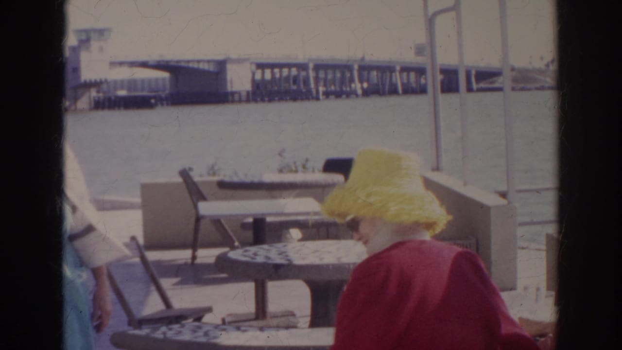 Woman in a yellow hat seated at a waterfront cafe table near empty chairs.