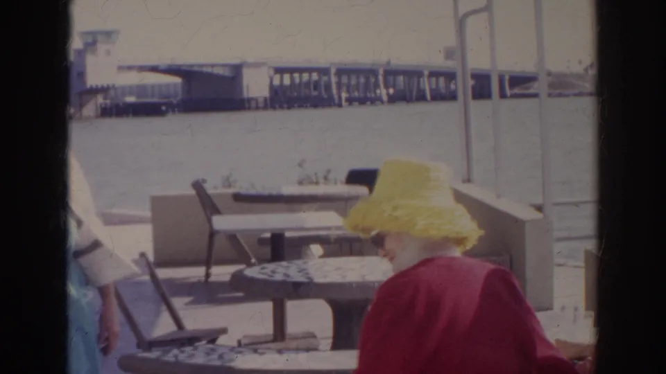 A woman in a bright yellow hat and red jacket sits at a Norfolk waterfront cafe table in this 1966 home-movie frame.