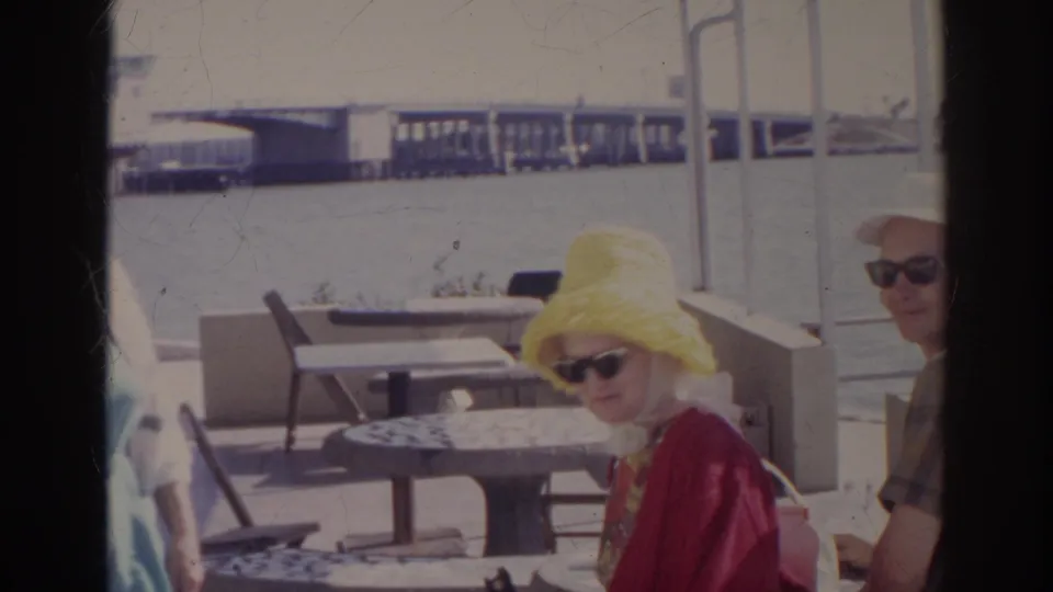 At the Norfolk waterfront cafe, a woman in a yellow hat and sunglasses sits beside another adult near stone tables and harbor water.