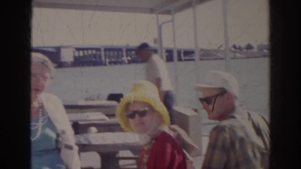 1966 Norfolk frame showing adults gathered at an outdoor waterfront cafe, including a woman in a yellow hat and a man in a white cap.