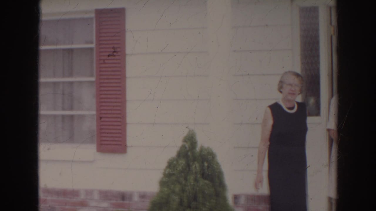 In supported 1966 Norfolk footage, a woman in a dark sleeveless dress stands near the doorway of a white clapboard house with red shutters.