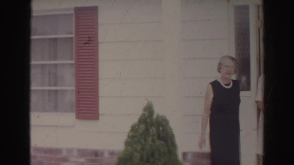 In supported 1966 Norfolk footage, a woman in a dark sleeveless dress stands near the doorway of a white clapboard house with red shutters.