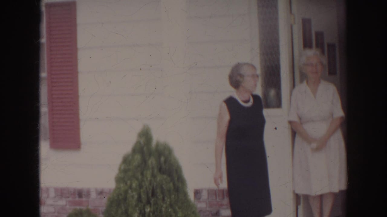 Two women stand outside a Norfolk residence in 1966, one in a dark dress and one in a light dress, near a front doorway and red shutter.