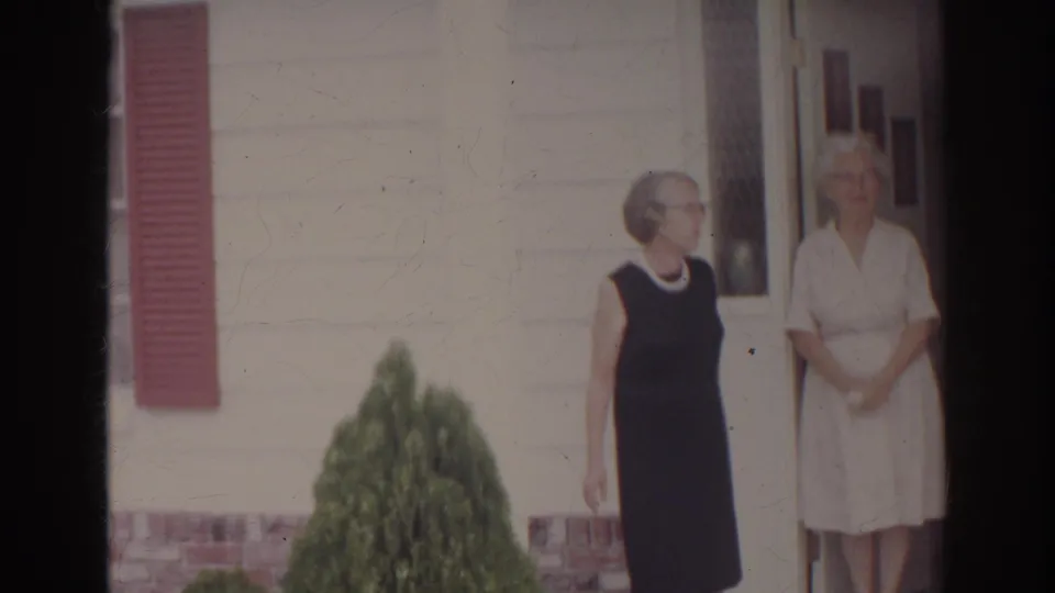 Two women stand outside a Norfolk residence in 1966, one in a dark dress and one in a light dress, near a front doorway and red shutter.