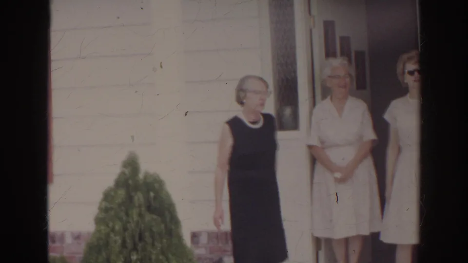 Three adult women in dresses stand outside a white clapboard house in supported 1966 Norfolk home-movie footage.