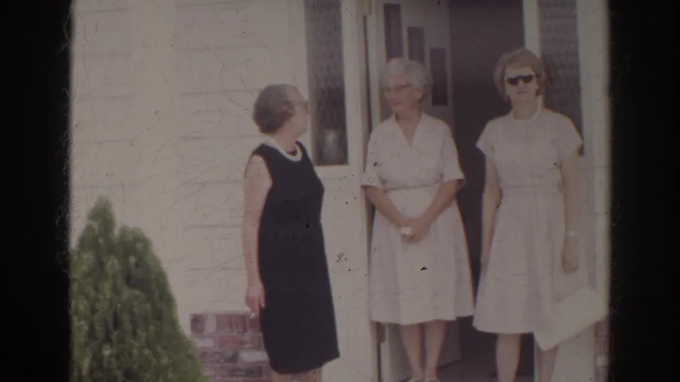Three women in dark and light dresses stand outside a Norfolk house in 1966, with red shutters, clapboard siding, and a shrub in view.