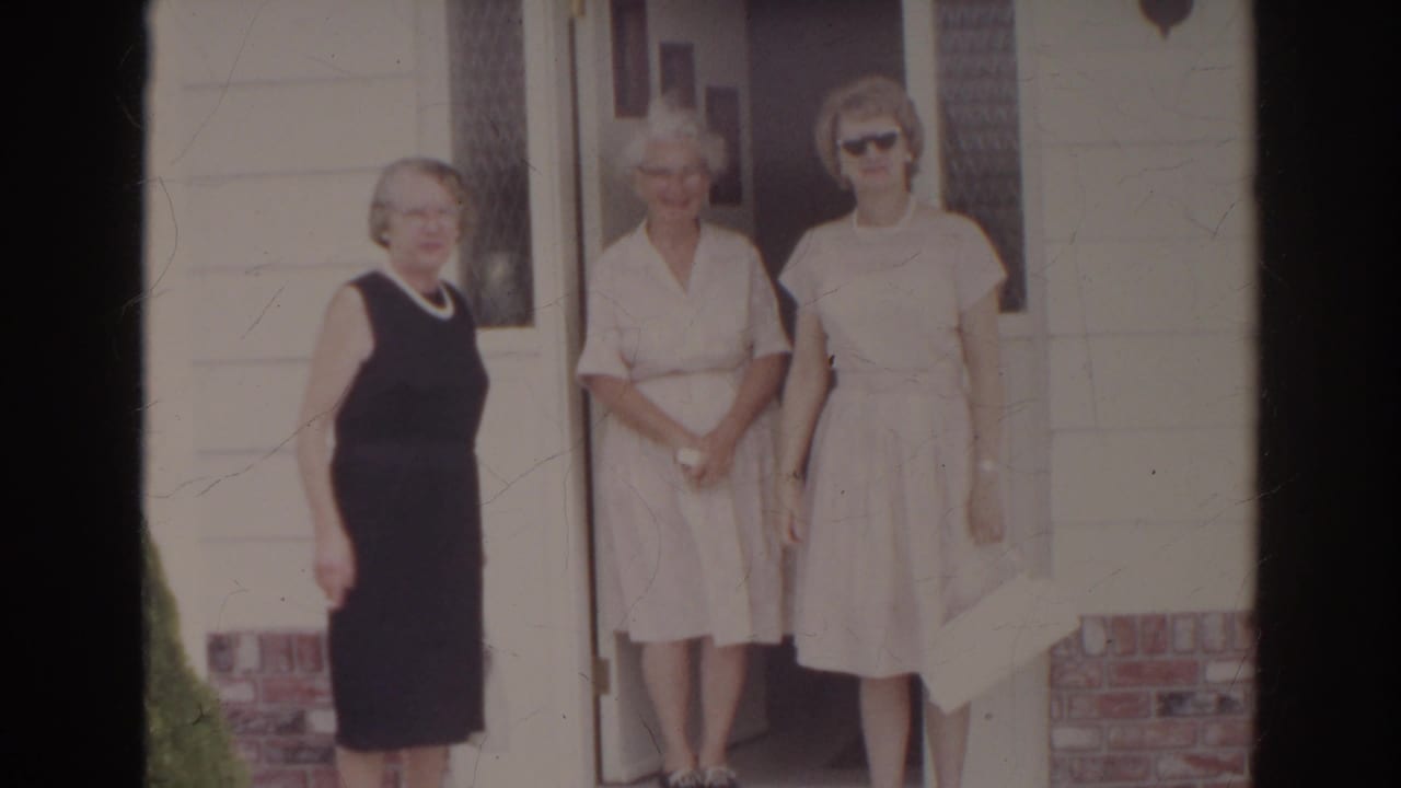 Three older women stand close together outside a Norfolk home, framed by siding, brick trim, and the open doorway.