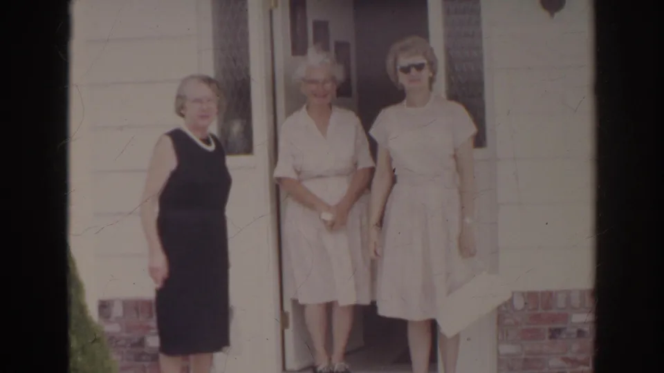 Three older women stand close together outside a Norfolk home, framed by siding, brick trim, and the open doorway.