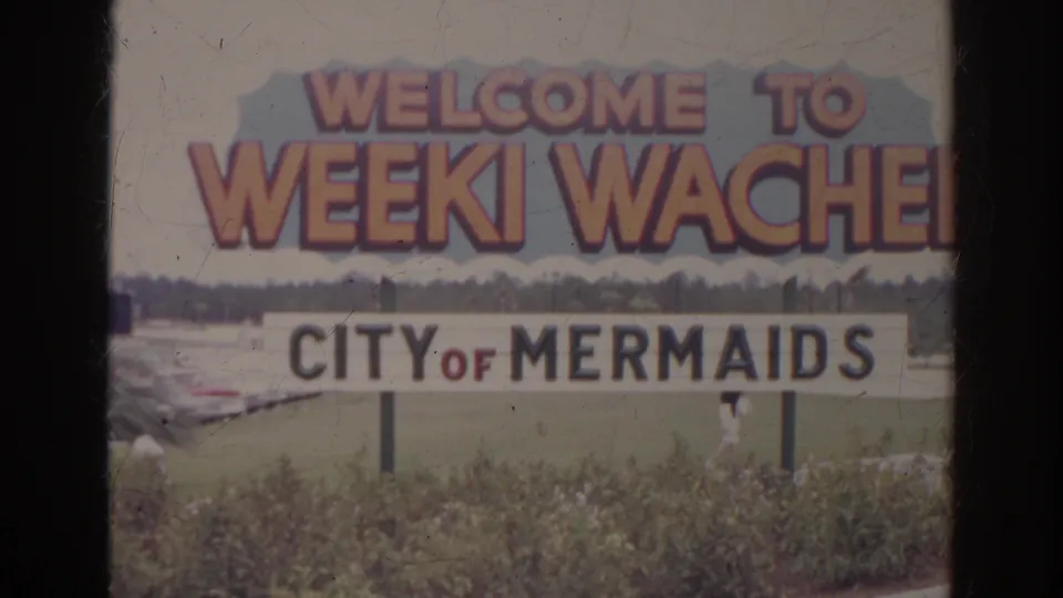 1966 8mm frame of a Weeki Wachee City of Mermaids welcome sign with pink lettering, greenery, and distant trees.
