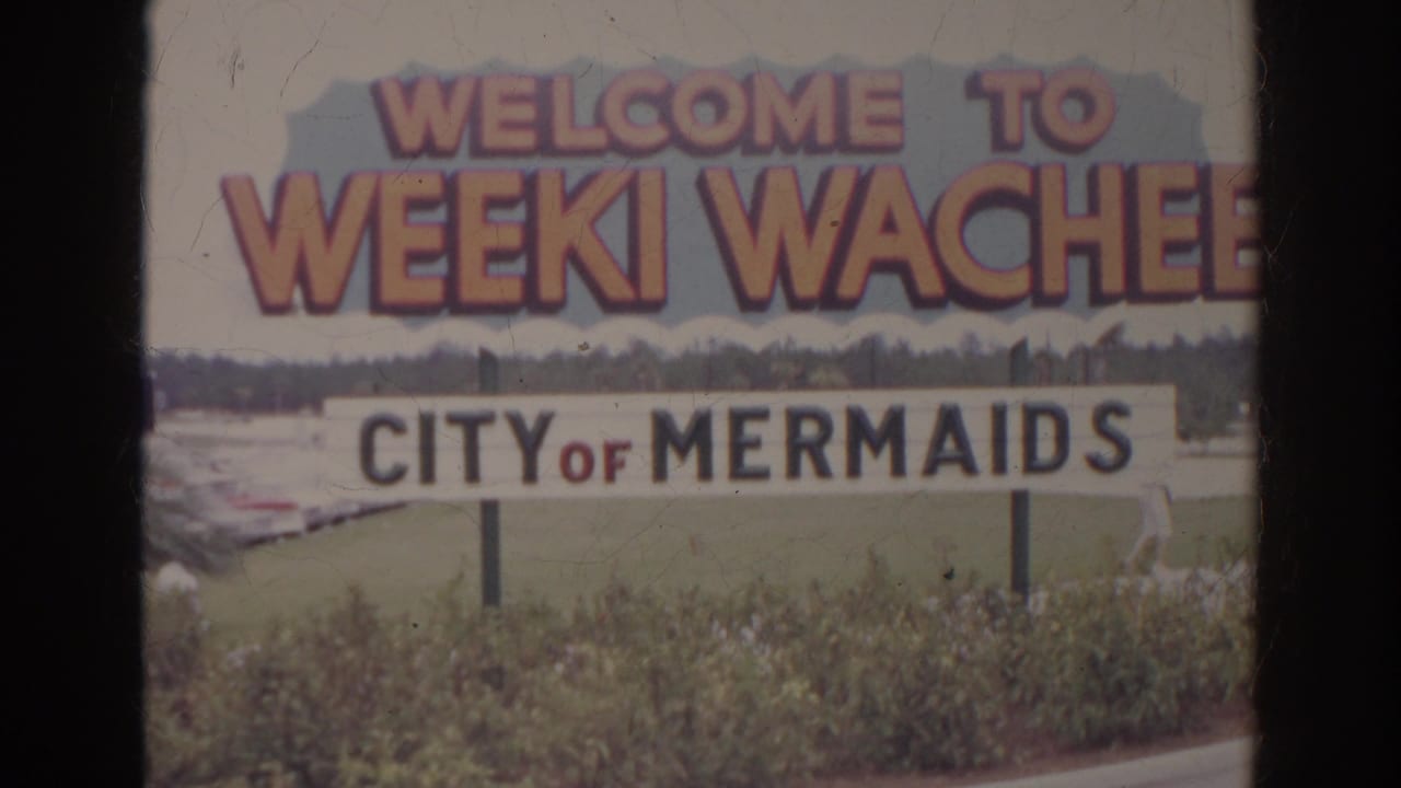 1966 8mm frame of the Weeki Wachee sign, with shrubs in front and a small person in white near the lower right area.