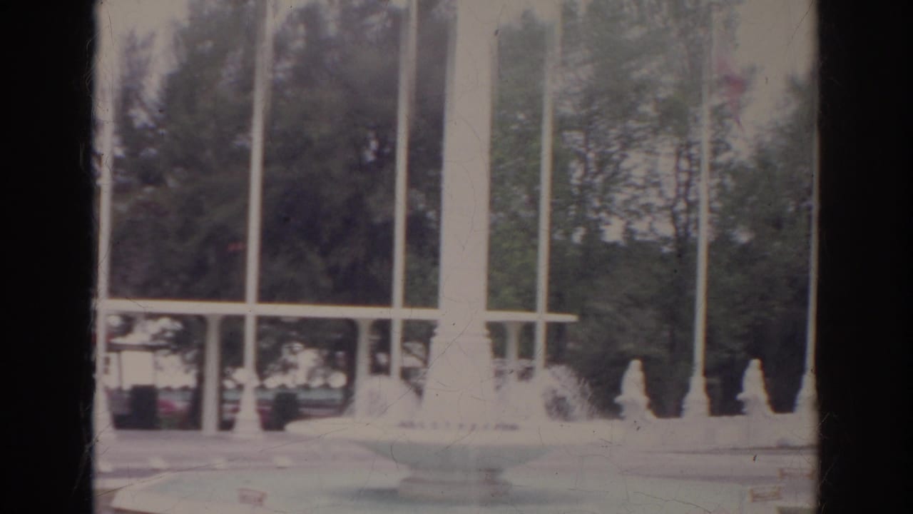 1966 8mm frame showing a fountain or decorative water feature with tall white columns, flagpoles, railing, and trees.