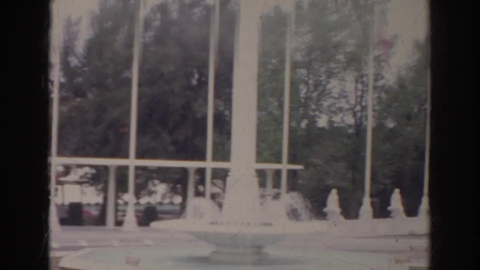 1966 8mm frame showing a fountain or decorative water feature with tall white columns, flagpoles, railing, and trees.