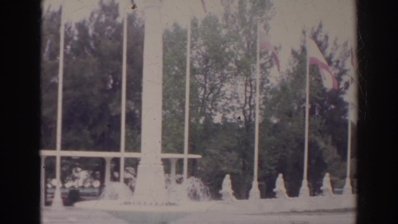1966 Norfolk, Virginia home-movie frame showing tall flagpoles, international flags, trees, and a white monument or fountain structure.