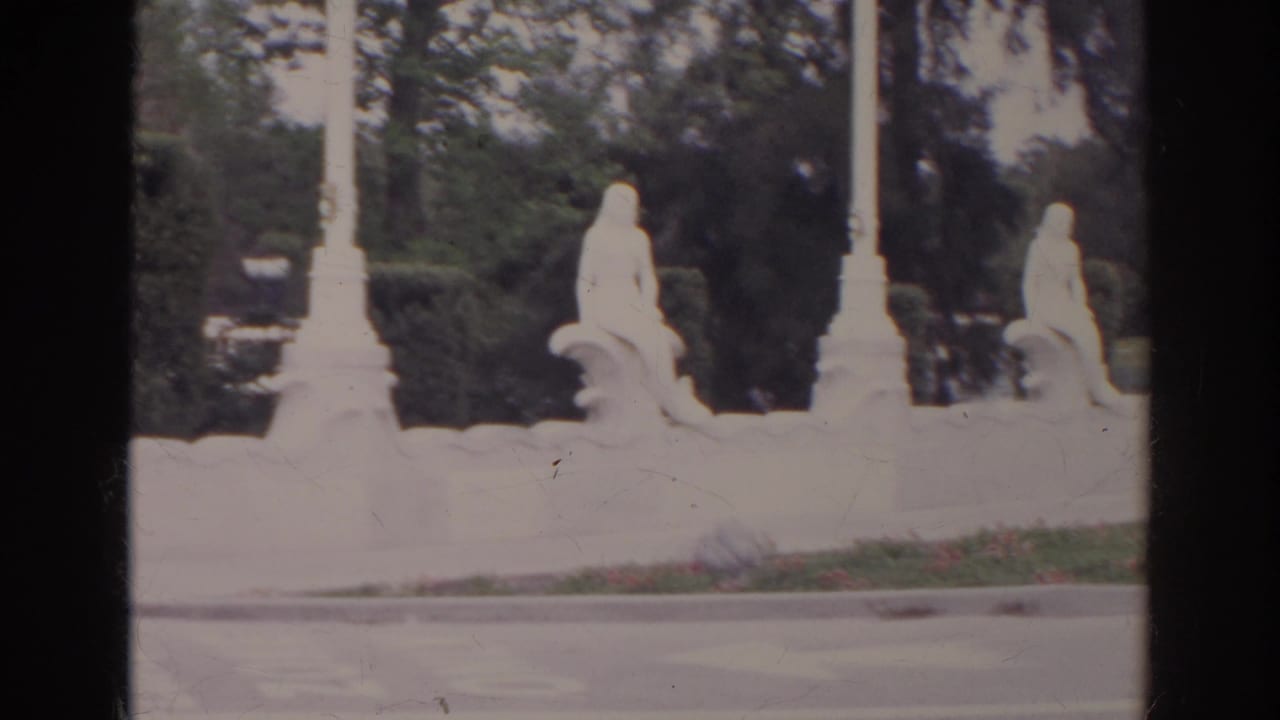 1966 Norfolk home-movie view of white fountain sculpture beside trimmed shrubs, flowers, curb, and roadway.
