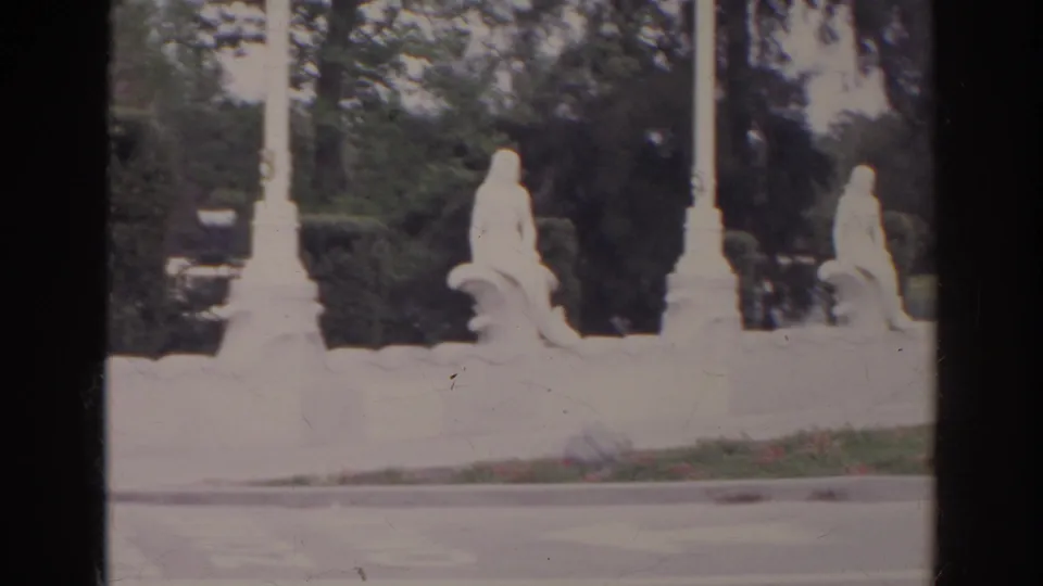 1966 Norfolk home-movie view of white fountain sculpture beside trimmed shrubs, flowers, curb, and roadway.